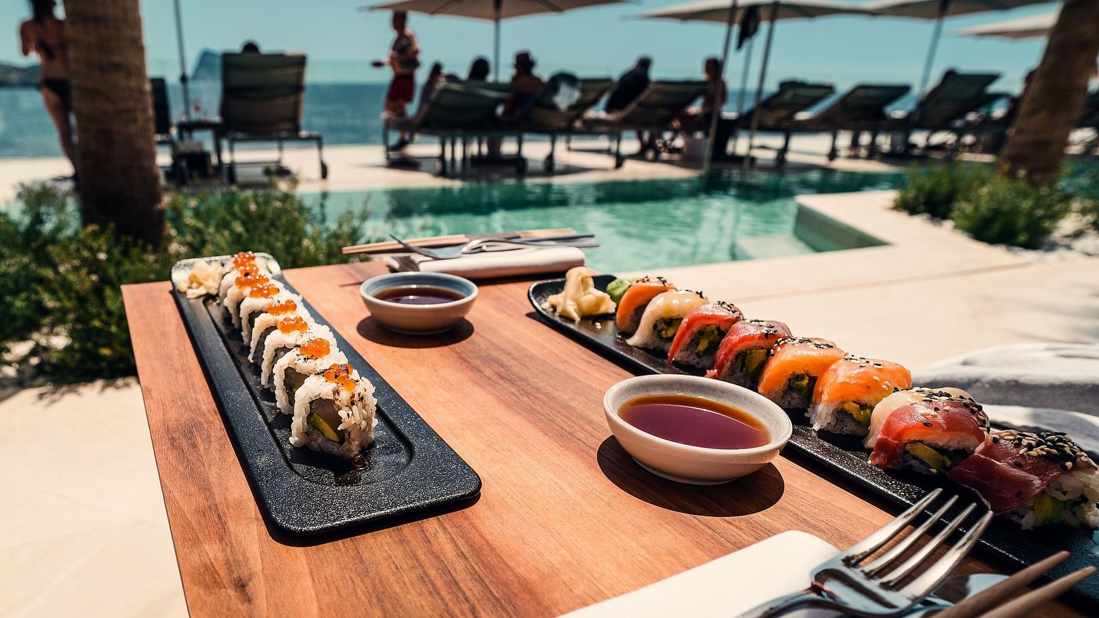 Sushi kept on a table with dips and cutlery neat the swimming pool with people resting on the loungers in the background