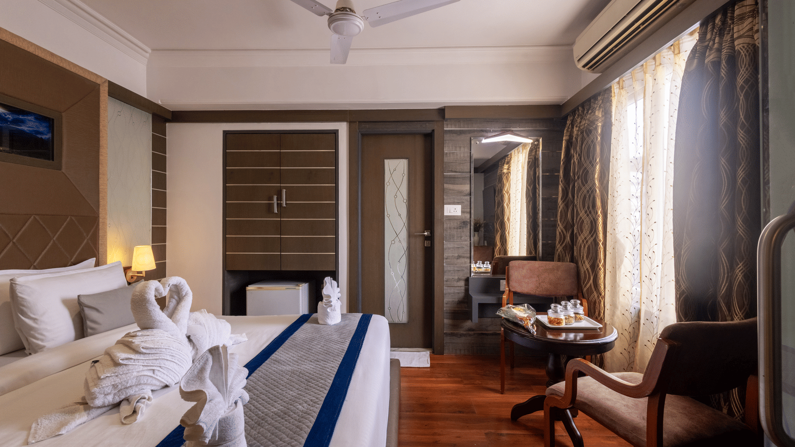 View of the bed reflected in a large mirror/television screen, highlighting the room's warm tones and wooden features at Pravasa Stay, Vadodara.