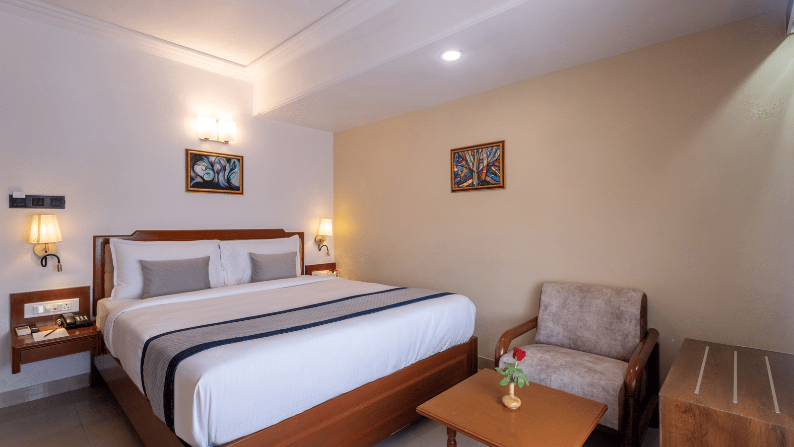 Close-up of a well-made hotel bed and a comfortable armchair with a side table, under ambient wall lights at Pravasa Stay, Vadodara.