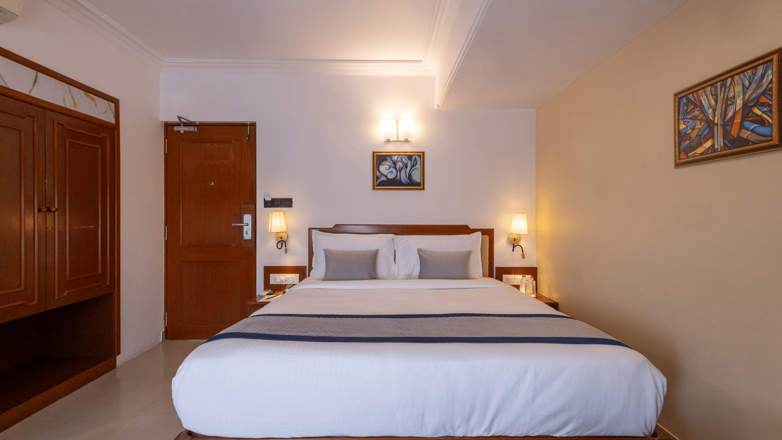 A view of the king-sized bed and headboard from the doorway, showing the wooden floor and warm bedroom lighting at Pravasa Stay, Vadodara.
