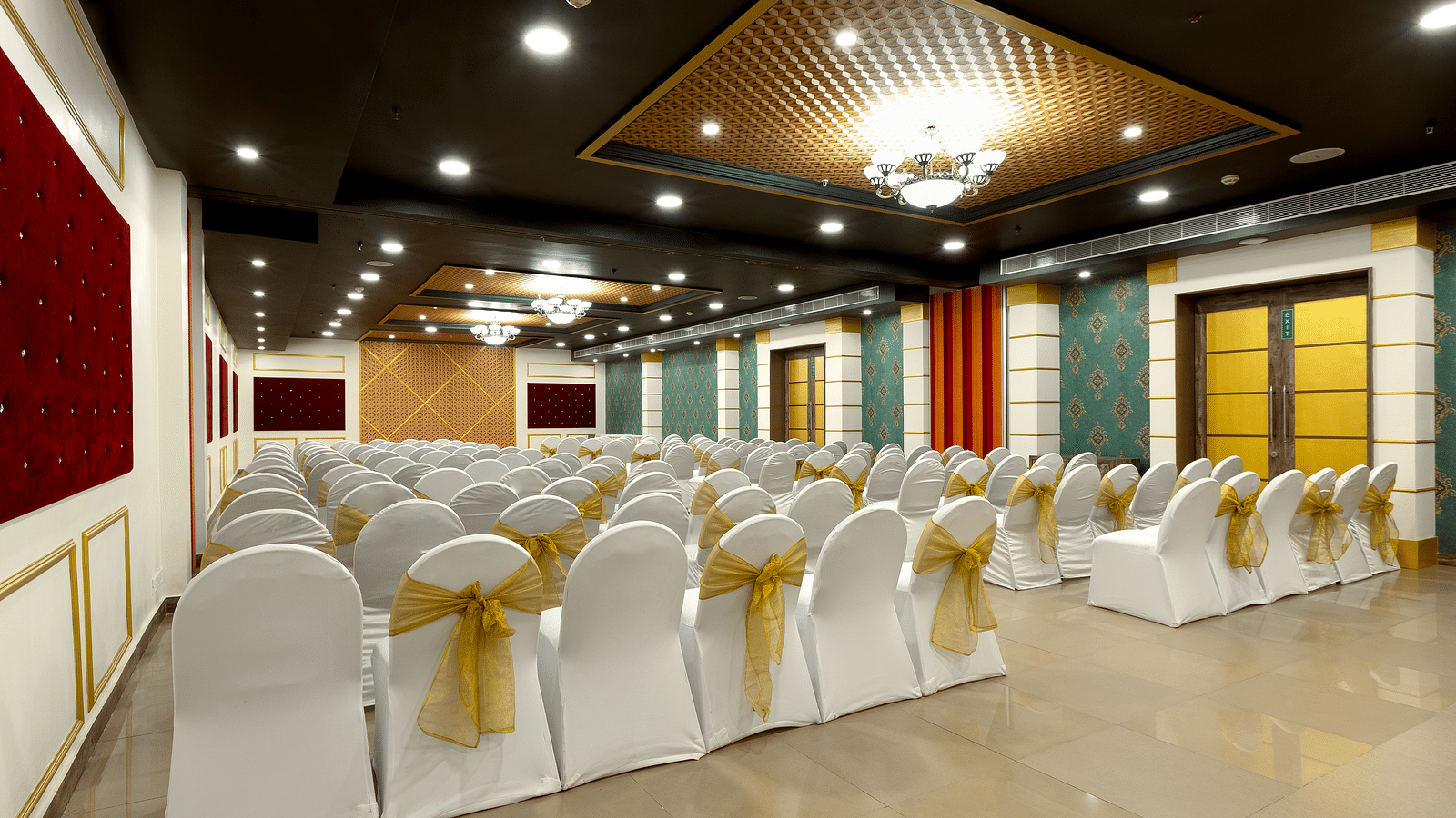 A spacious banquet hall with chairs covered in white cloths and gold  covers. The hall has a modern design with dark wood accents and a crystal chandelier.