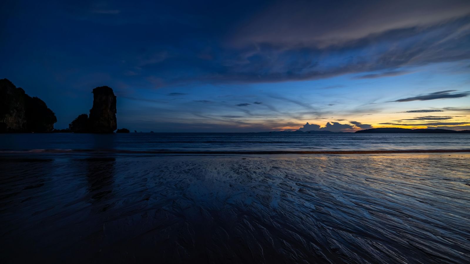 A dramatic, dark shot of a beach at dusk with a solitary rock formation in the distance.