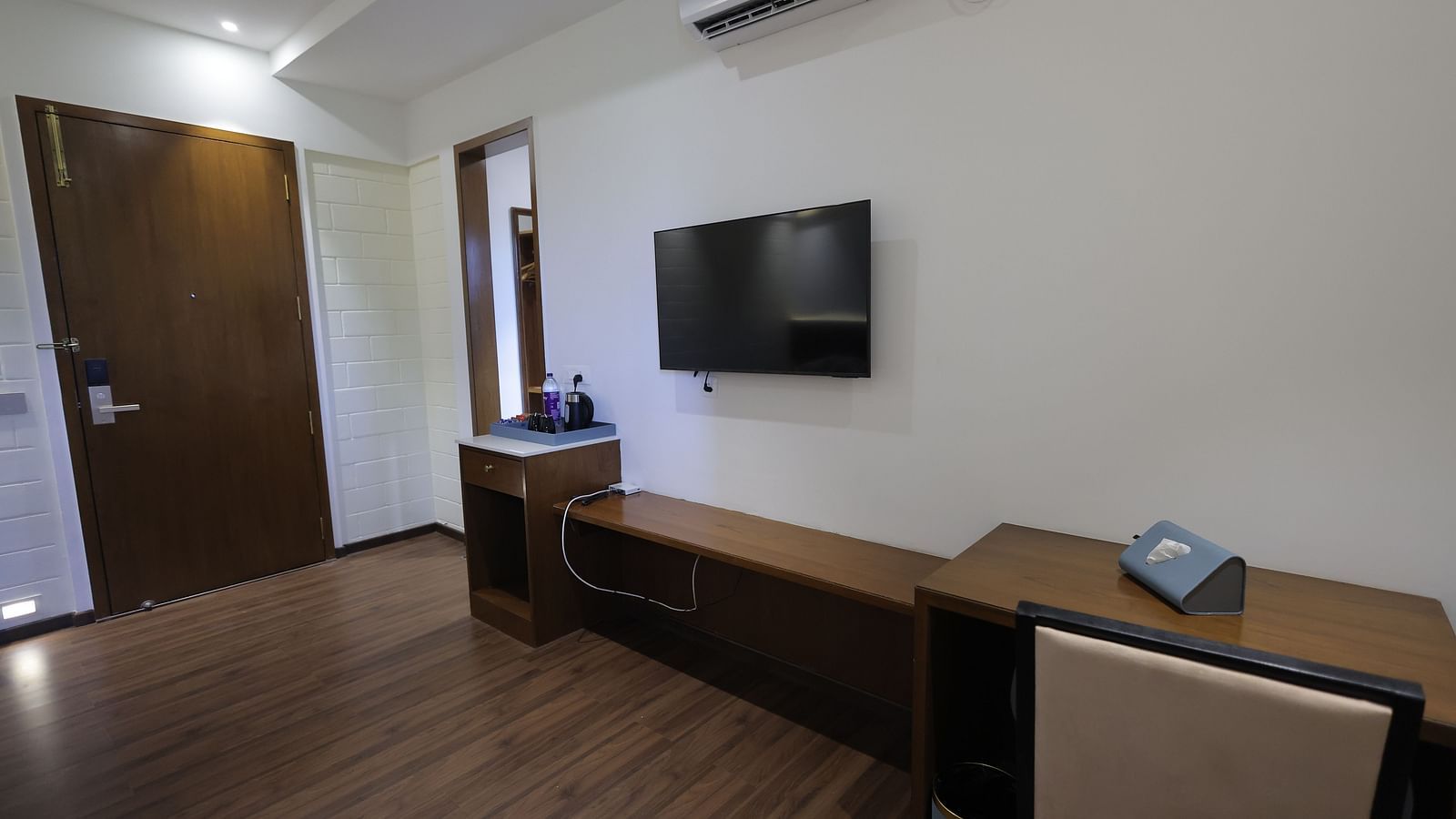 Room with wall-mounted TV, desk, chair, and wooden flooring at Trishvam Symphony, Krishnagiri.