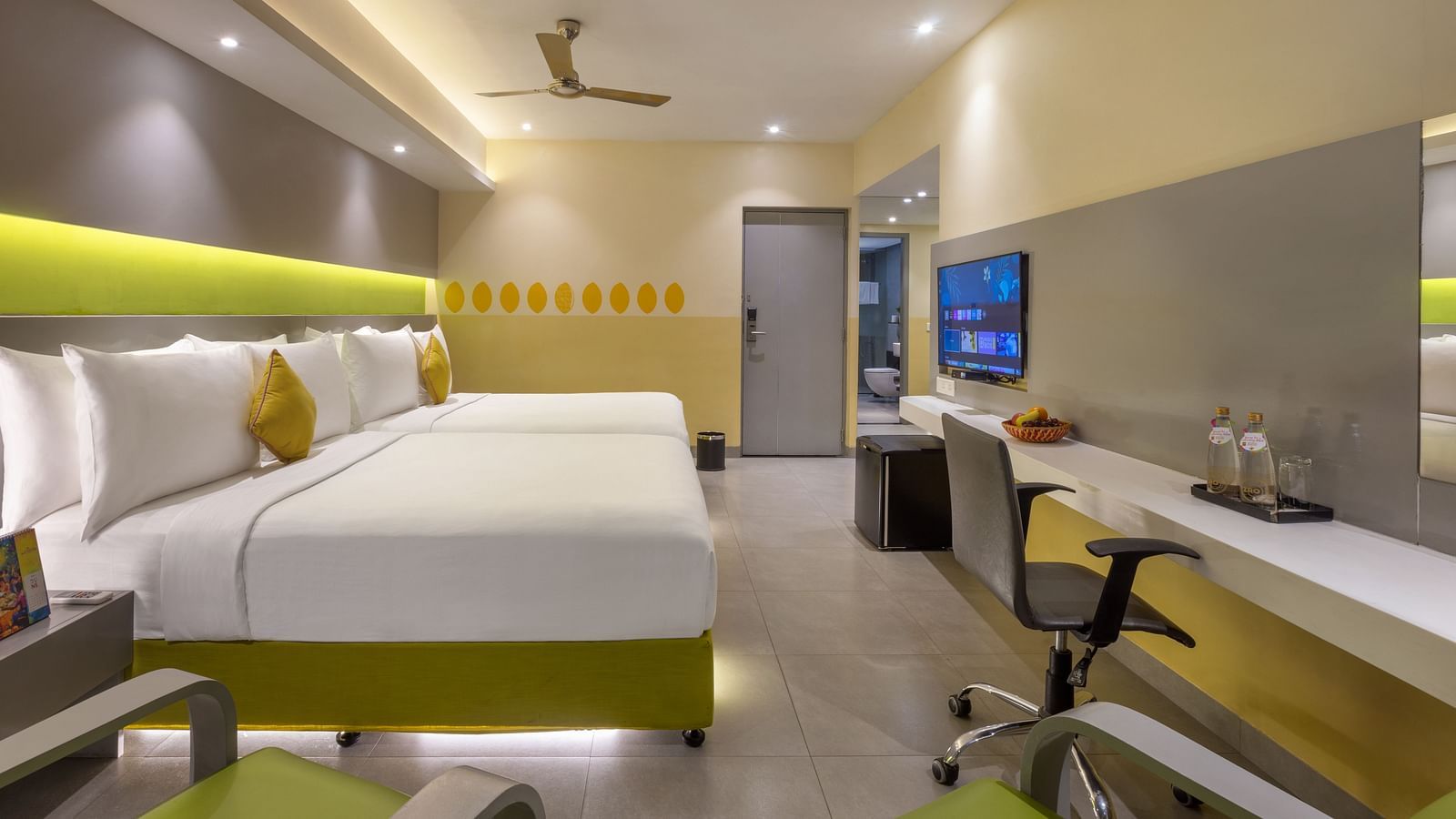Spacious hotel room at Zibe Luxe Hyderabad by GRT Hotels with two beds, green armchairs, a desk with a TV, and a green backlit panel above the beds.