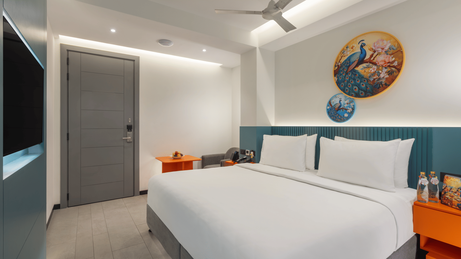 A room at Zibe Luxe Hyderabad by GRT Hotels featuring a bed, 2 wall paintings,  a wall-mounted television facing the bed, and a couch and a coffee table in the corner.