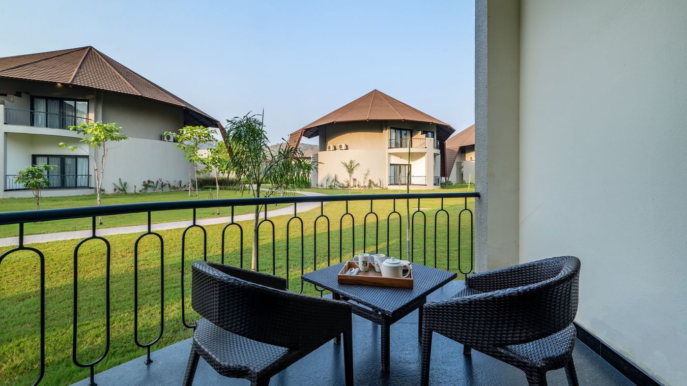An image of a balcony with two wooden chairs and a tea table from where other villas and lawn can be seen - Ananta Spa and Resort, Ajabgarh