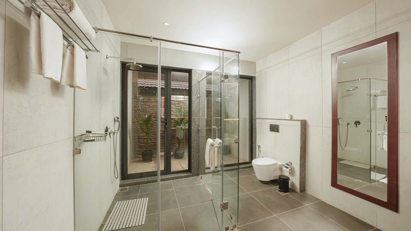 Bathroom of Lagoon Villa at Ananta Spa & Reosrt, Ajabgarh, featuring a glass partition, bathroom fittings, a large wall-mounted mirror, and a western commode.