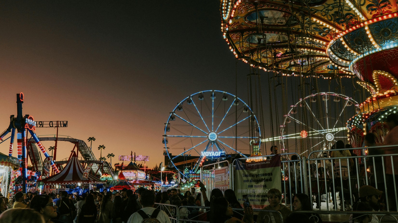 A bustling carnival is captured at night, showcasing illuminated rides including a prominent Ferris wheel | Dive Festival Havelock Island