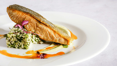 A cooked salmon fillet is presented on a white plate with a green garnish and an orange sauce drizzle.