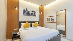 A bright hotel bedroom with a large bed, yellow pillows, a patterned grey headboard, and a framed piece of artwork depicting a building on the wall - Pride Premier Dehradun