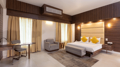 A modern hotel bedroom with a large bed, dark wood panelling above the headboard, and a sitting area with an armchair and table - Pride Premier Dehradun