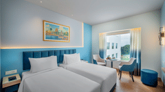 Modern twin hotel room with two beds, a blue padded headboard, a window overlooking a bright outdoor view, and a small table with two chairs.