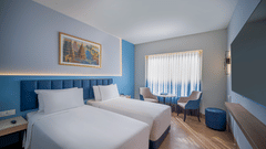 A hotel room with two single beds, blue accent walls, and a window with curtains, suggesting a comfortable twin accommodation | Standard Twin Room | Sarovar Portico, Sriperumbudur