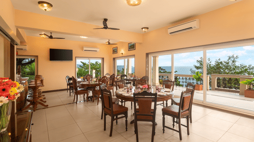 A dining area with vintage style furniture and a deck with the view of the ocean and lush green trees - Highland Sarovar Portico, Sindhudurg