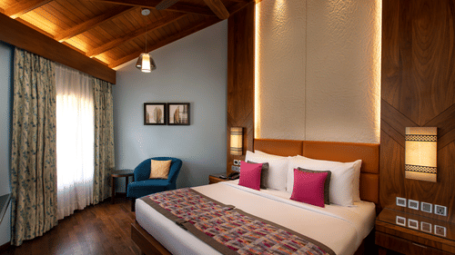 An overview of the Presidential Suite with a king bed, white and red pillows, a blue chair, paintings on the wall, accent lights above the headboard, and side tables and lamps next to the bed at Kenilworth Resort & Spa, Goa, the ideal place for a stay in G