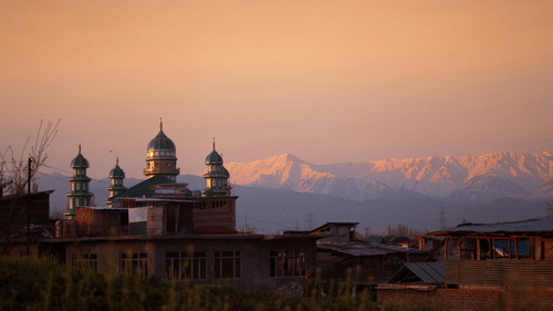 Aishmuqam Shrine seen with pointed domes, and nearby buildings in the foreground while huge expansive snow-capped hills cover the backdrop of the shrine.