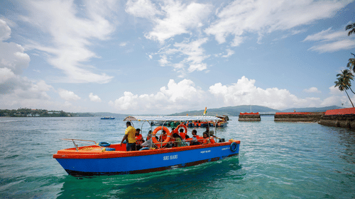 A vibrant blue and orange boat full of people is floating on the clear blue water of a bay under a bright sky.