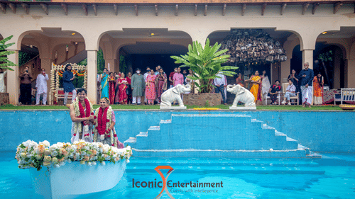 A couple in traditional attire is on a flower-adorned boat in a pool at Tijara Fort-Palace - 19th Century, Alwar, with guests standing by the poolside.