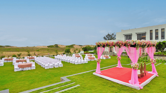 An outdoor wedding venue setup on a lush green lawn, featuring rows of chairs facing a floral archway, with a large, modern white building and palm trees in the background under the sky at Pramod Hotels & Resorts.