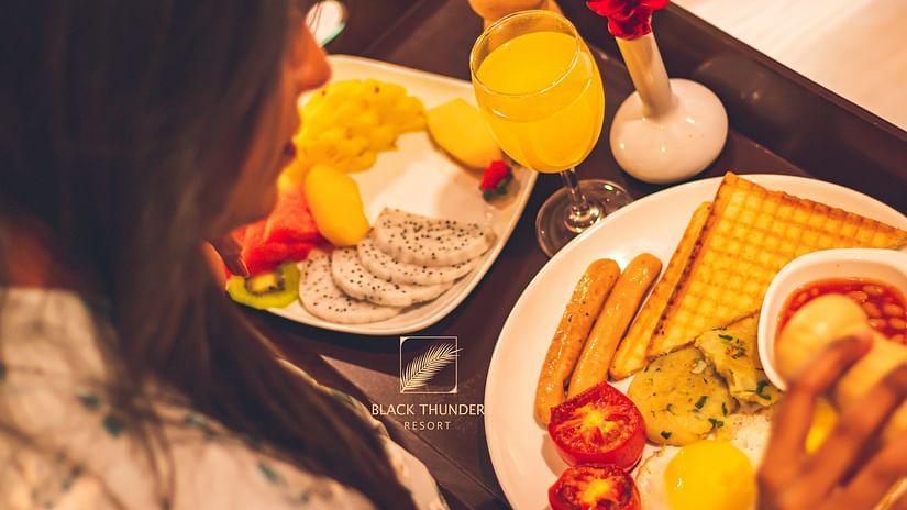 A close-up view of a person’s breakfast plate with toast, fruits, and a glass of orange juice, placed on a wooden table - Black Thunder, Coimbatore