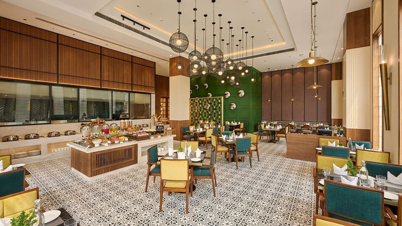 A spacious hotel restaurant features tables with colourful chairs set on a patterned floor, with a buffet area and modern pendant lighting creating a welcoming atmosphere. - Iris Sarovar Portico, Sector 85, Gurugram