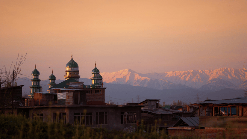 Aishmuqam Shrine seen with pointed domes, and nearby buildings in the foreground while huge expansive snow-capped hills cover the backdrop of the shrine.