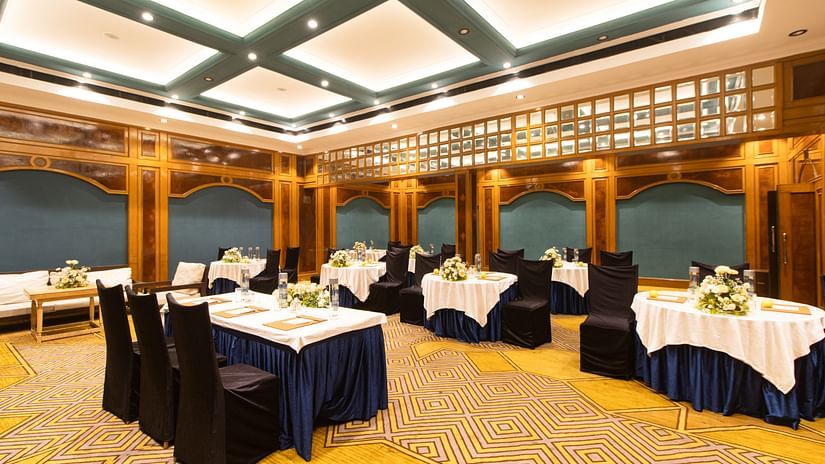Grand banquet hall with geometric carpet, wood paneling, round tables with blue skirts, and tastefully arranged flowers for a formal event. - Polo Floatel, Kolkata
