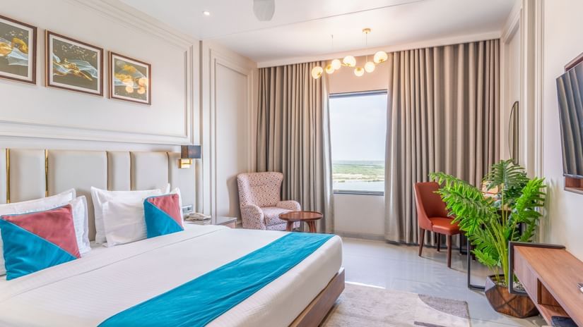 Premium Room at Pride Comfort Dwarka showcasing a double size bed, a bedside table with lamp, a sofa chair with centre table, a work station, a flat TV screen, and a flower pot.