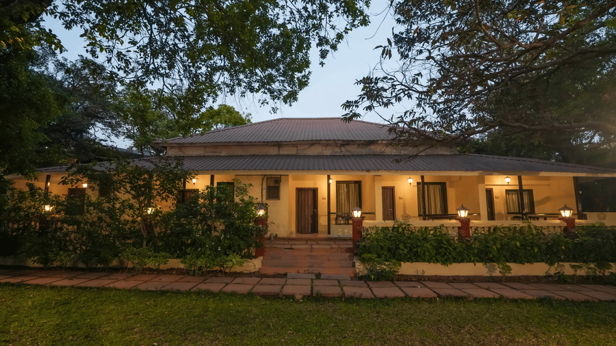 Exterior view of a long, single-storey resort building with a porch, surrounded by a lawn and trees at Adamo The Village.