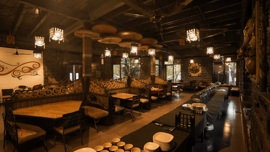 A wide view of dining area of Adamo The Village featuring rustic decor, ceiling lights, and wooden furniture arranged neatly in rows.