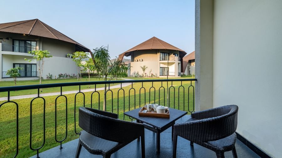 An image of a balcony with two wooden chairs and a tea table from where other villas and lawn can be seen - Ananta Spa and Resort, Ajabgarh