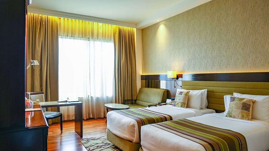 A bright and welcoming deluxe room with twin beds, a comfortable seating area, wooden flooring, and soft curtains allowing natural light - Balaji Sarovar Premiere, Solapur
