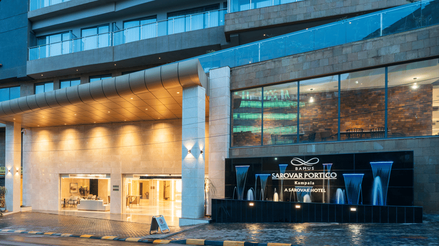 Facade of Bamus Sarovar Portico seen with a well-lit portico. Signage of the hotel is also seen with a water fountain on a side.