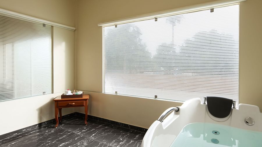 Bathroom of Signature Suite featuring a jacuzzi by a window and a small wooden table with toiletries at DGV Resort by Treat, Silvassa.