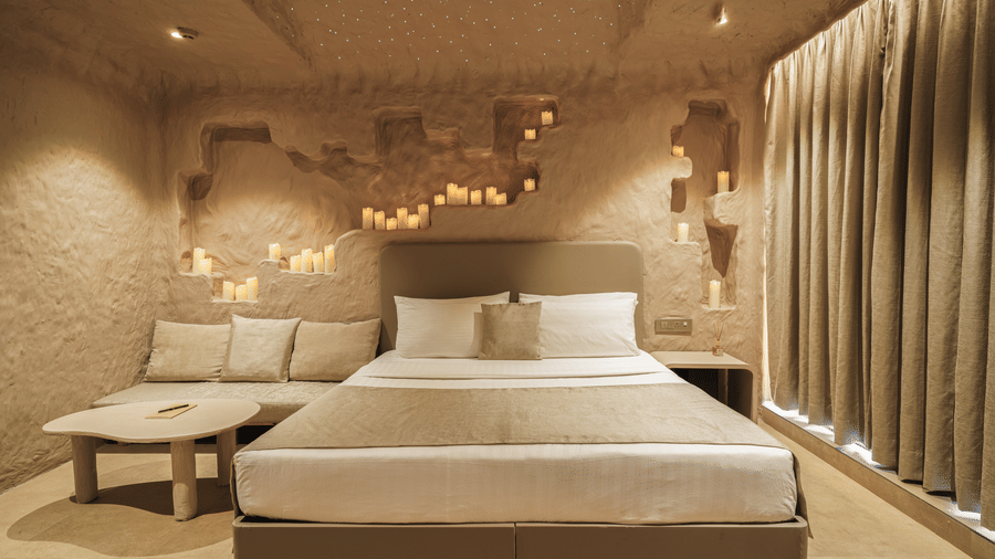 Cave Room at Dragonfly Hotel – The Art Hotel with textured stone?like walls and ceiling, large bed with white bedding, cushioned bench, recessed lighting, and warm neutral decor.