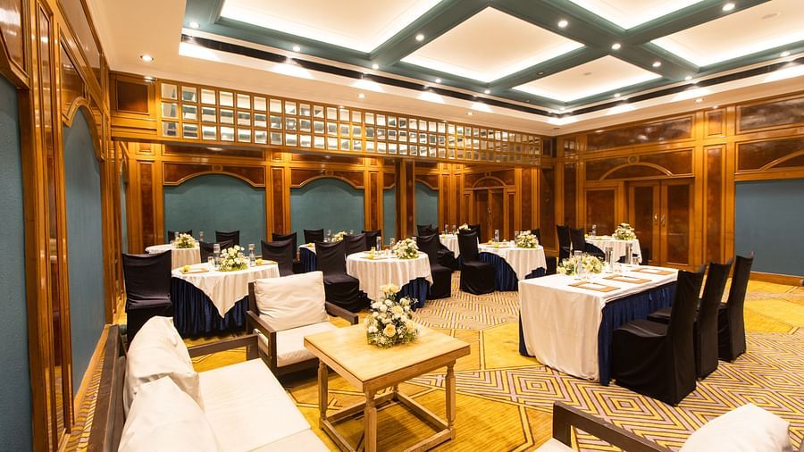 Grand banquet hall with geometric carpet, wood paneling, round tables with blue skirts, and tastefully arranged flowers for a formal event.