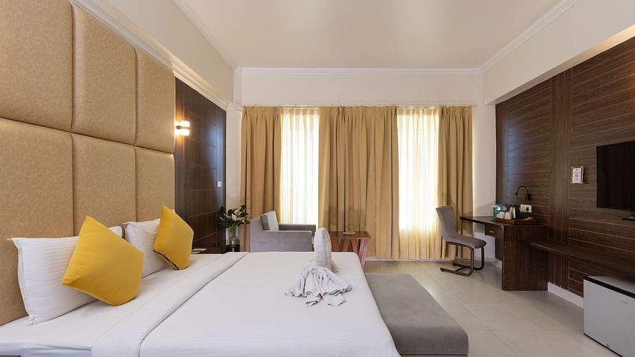 A hotel bedroom with a large bed, yellow accent pillows, a padded headboard, and two white folded towels on the bed - Pride Premier Dehradun