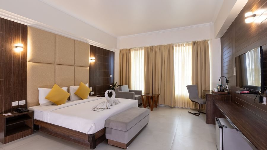 A wide shot of a hotel bedroom with a large bed featuring white swan-shaped towel decorations, a padded headboard, a television, and a sitting bench - Pride Premier Dehradun