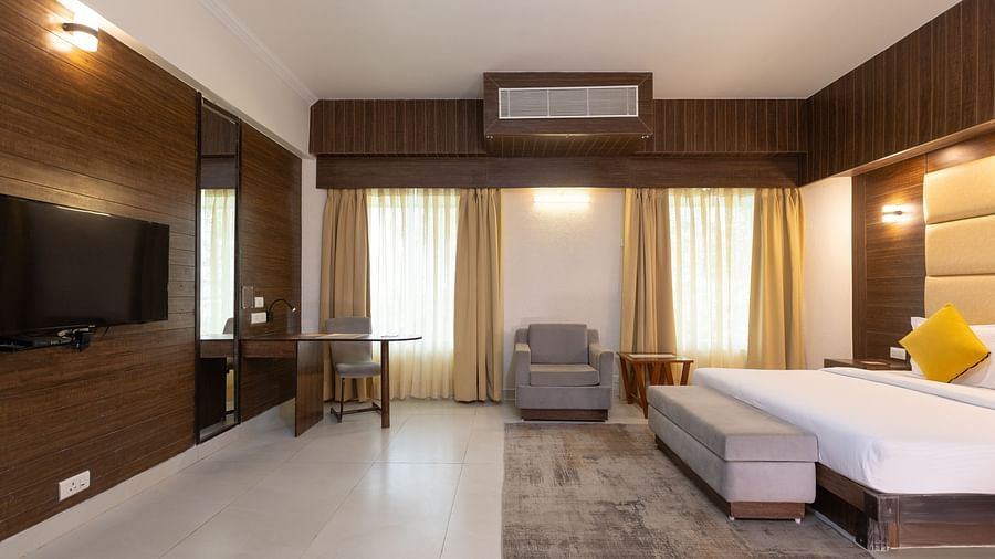 A wide view of a modern hotel bedroom, showing a large bed, a dark wood feature wall with a mounted television, a desk, and a small sofa - Pride Premier Dehradun