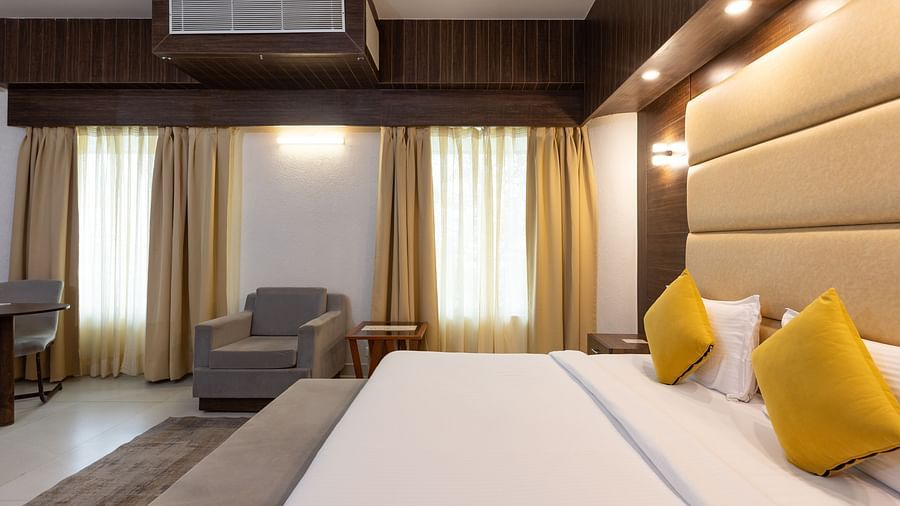 A contemporary hotel bedroom with a large bed dressed in white linen and yellow pillows, a light-coloured padded headboard, and an armchair in the corner - Pride Premier Dehradun