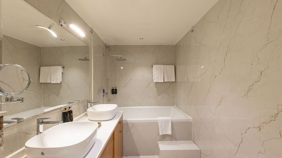 A spacious, bright bathroom with two oval basins on a light counter, a large mirror, a bathtub, and a shower area - Pride Premier Solitaire Dehradun