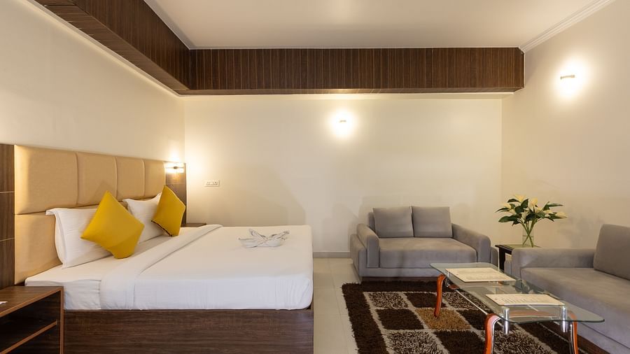 A hotel bedroom with a large bed with yellow pillows, a comfortable sofa, and a coffee table, all set on a light-coloured floor - Pride Premier Dehradun