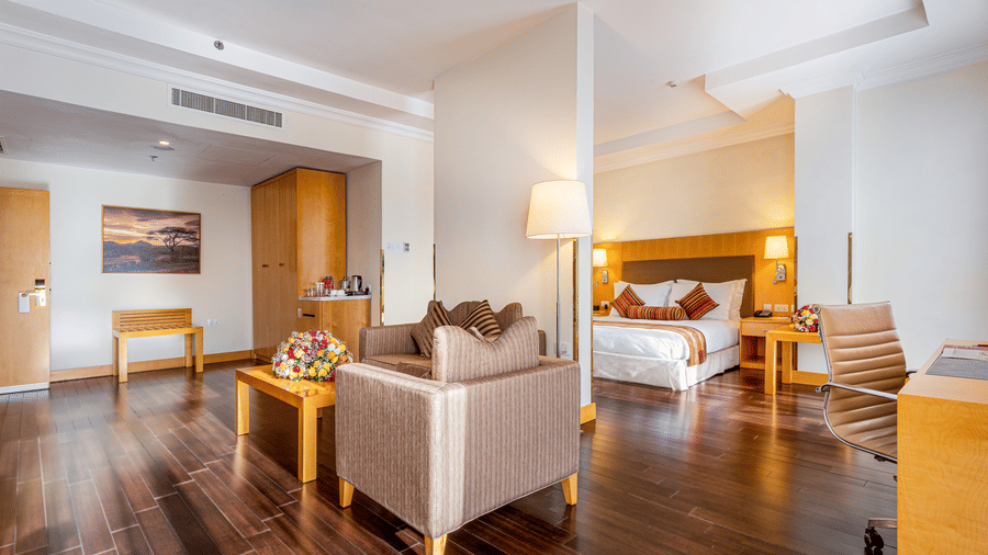 A suite with a bed, seating area, desk, chairs, tables, lamps, wooden floor, wall art, wardrobe, and a tea and coffee setup at Ramada by Wyndham Addis, Addis Ababa.