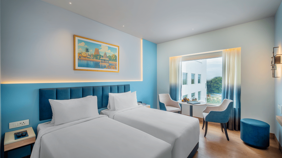 Modern twin hotel room with two beds, a blue padded headboard, a window overlooking a bright outdoor view, and a small table with two chairs.