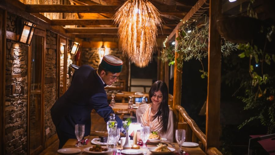 Outdoor dining experience under a glowing hanging lamp with guests enjoying a meal within a wooden pergola surrounded by greenery, highlighting the sophisticated and intimate culinary atmosphere at The Manor Sports & Wellness Hotel in Shimla.