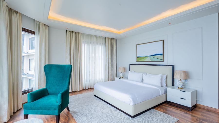 Bright, airy bedroom decorated in white tones with accent furniture and expansive windows, reflecting the stylish and contemporary interiors found at The Manor Sports & Wellness Hotel in Shimla.
