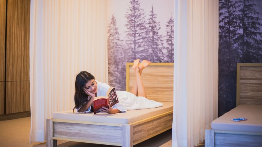 A women lying and reading book in a  cosy, alpine-themed hotel room at The Manor Sports & Wellness Hotel in Shimla.