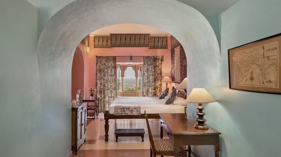 Hotel suite with a view through an arched opening into a bedroom with a bed, patterned curtains, and a balcony, and a hallway with a desk and artwork at Tijara Fort-Palace, Alwar.