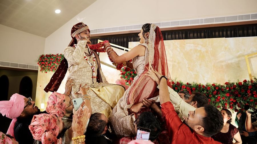 An image of a wedding celebration with the bride and groom being lifted in the air by family and friends, and the bride, holding a garland, is trying to put it on the groom's head