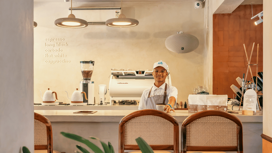 A staff member in white uniform and cap standing behind a clean cafe counter at The Bakehouse with coffee equipment wooden cabinetry rattan chairs and warm overhead lighting in a bright setting.
